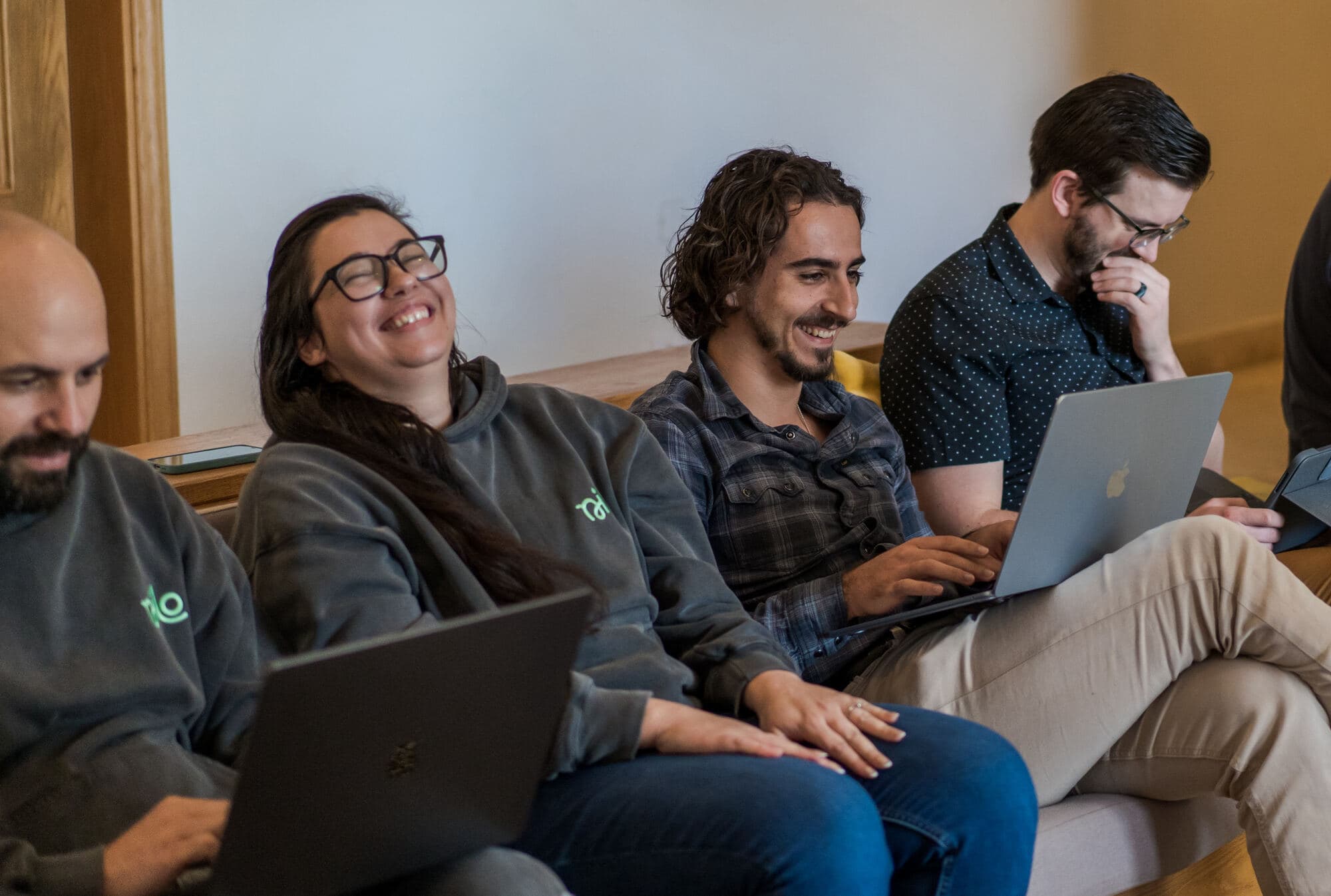Four Nilo teammates laughing together on a bench, each with a laptop open, during an offsite.